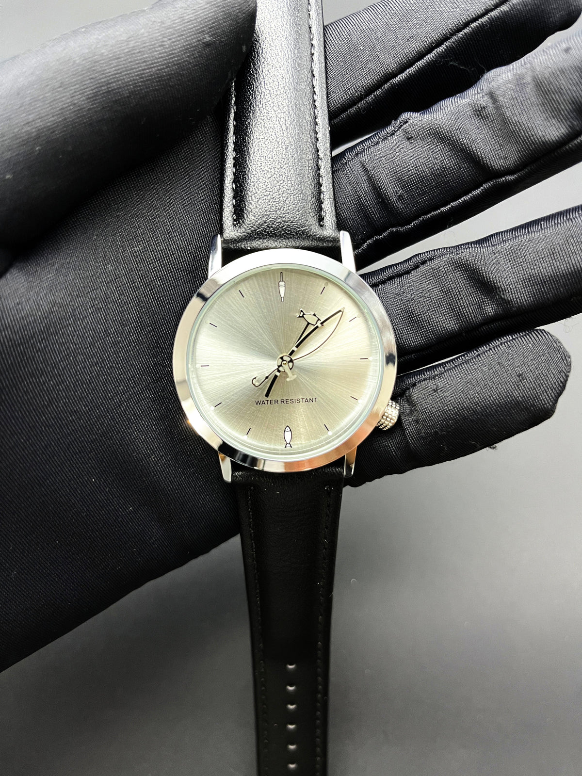 CHOOSZ Lucky Cast watch held in a black-gloved hand, showing its sunburst dial, fishing-themed hands and black leather strap
