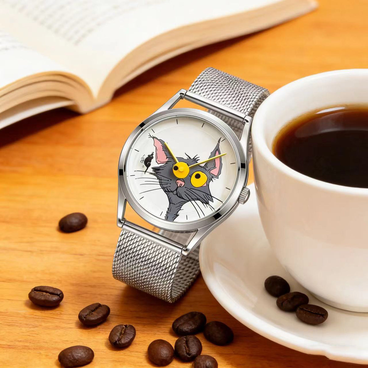 Chase Me, Kitty! cat dial watch with silver mesh strap beside a coffee cup, coffee beans and an open book on a wooden table