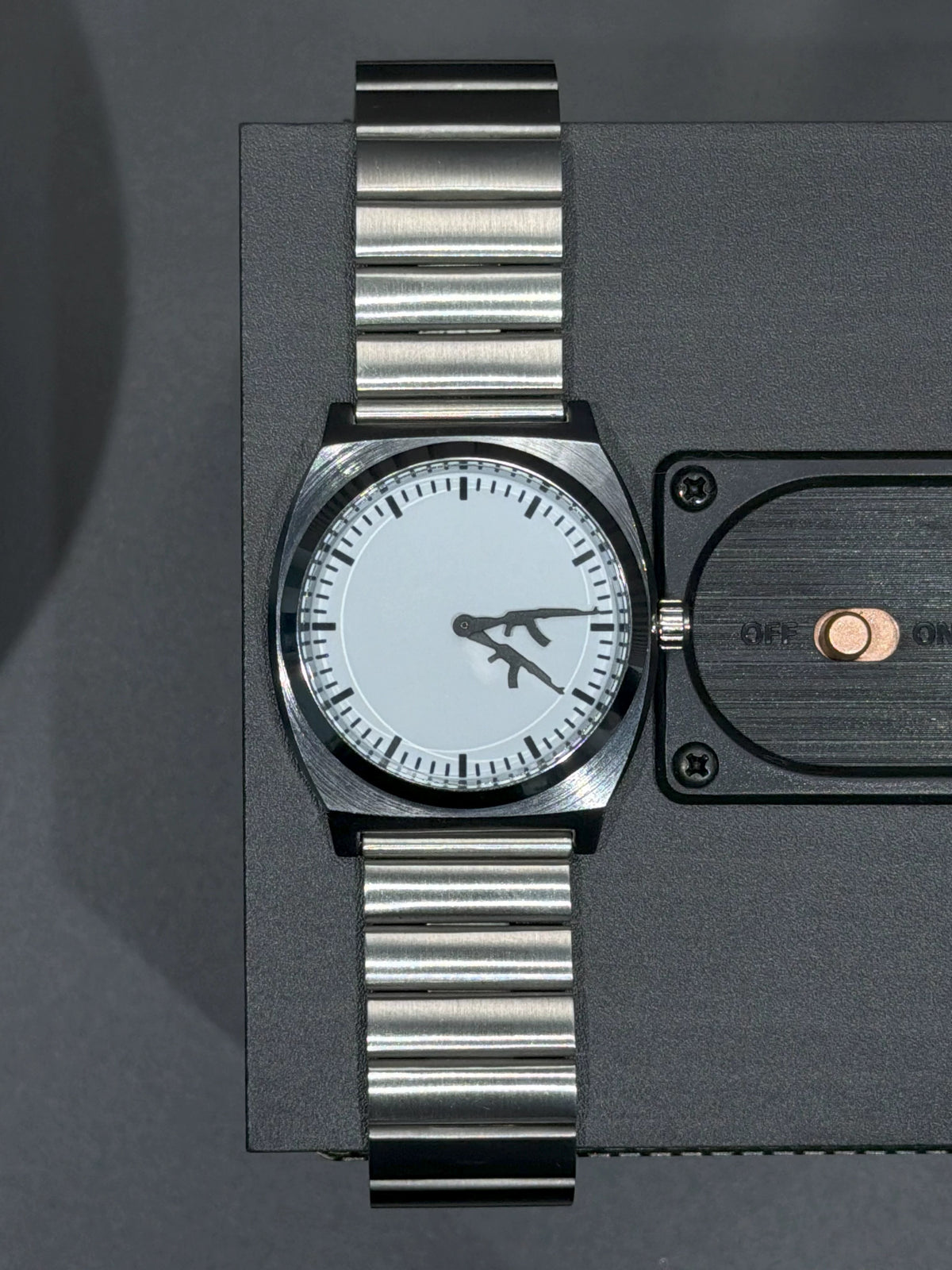 Top view of CHOOSZ Arcade Loadout watch on a black speaker, showing white dial, gun-shaped hands and silver steel bracelet