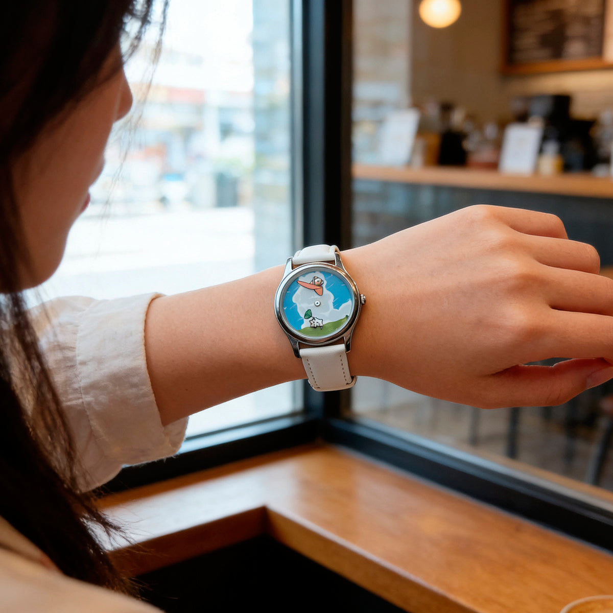 CHOOSZ Cloud Companions watch with blue illustrated dial and white leather strap worn on the wrist beside a cafe window