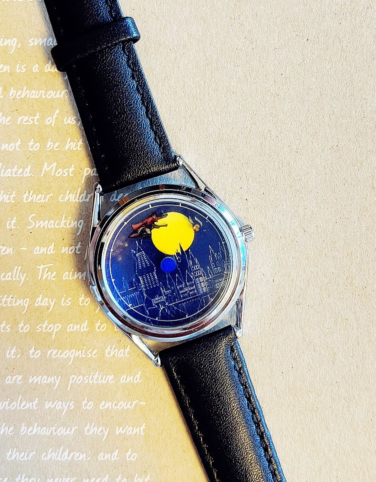 CHOOSZ The Golden Chase watch with blue castle-and-moon dial, silver case and black leather strap resting diagonally on a printed page