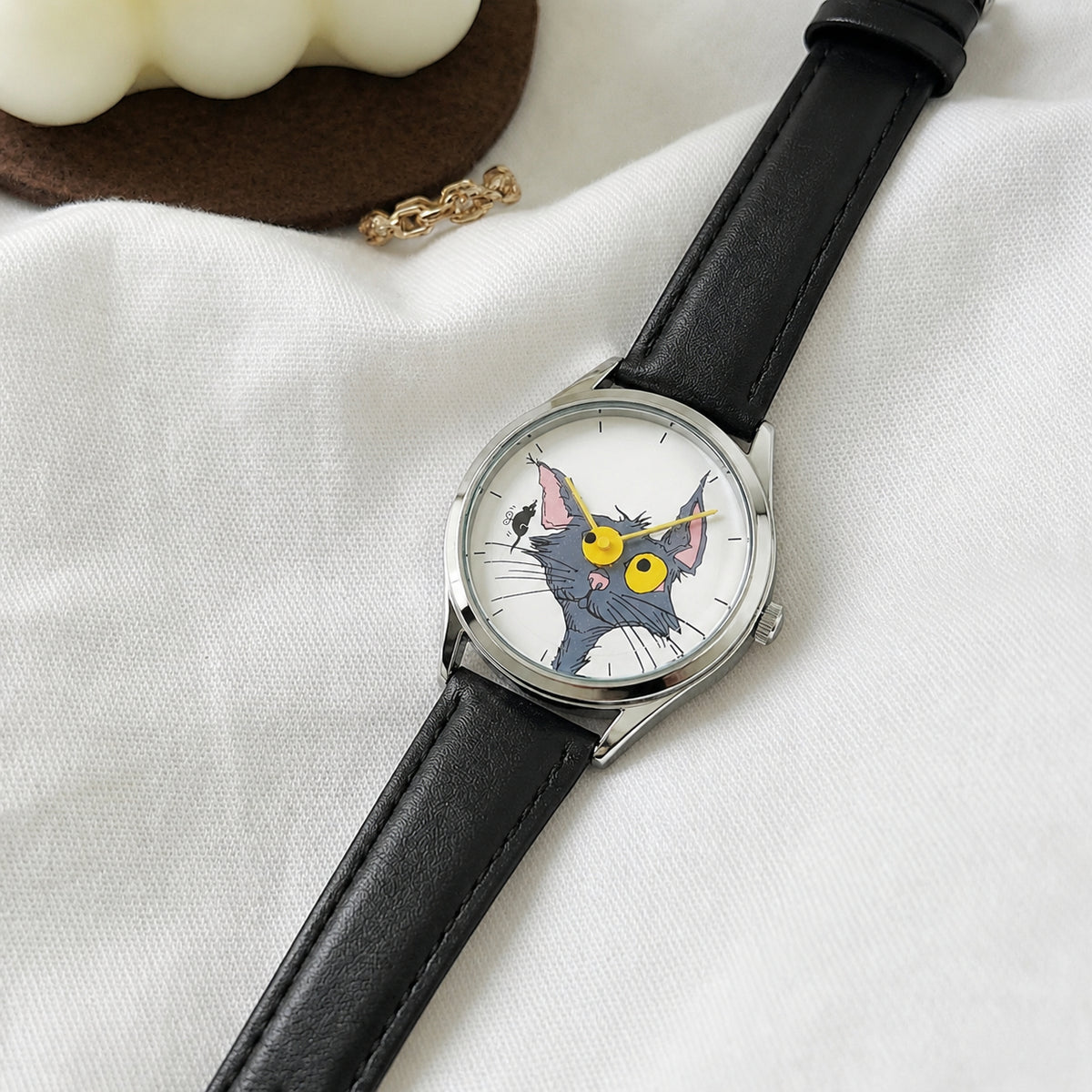 Chase Me, Kitty! cat-and-mouse dial watch with black leather strap laid diagonally on white fabric