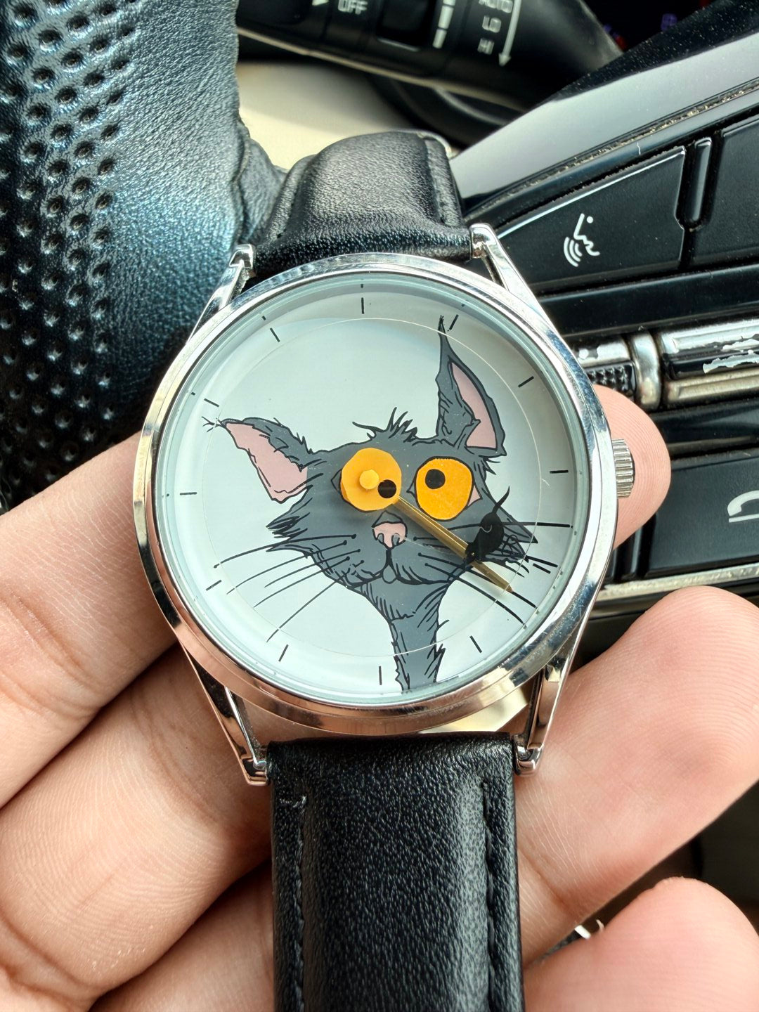 Chase Me, Kitty! cat-and-mouse dial watch with black leather strap held in front of a car steering wheel